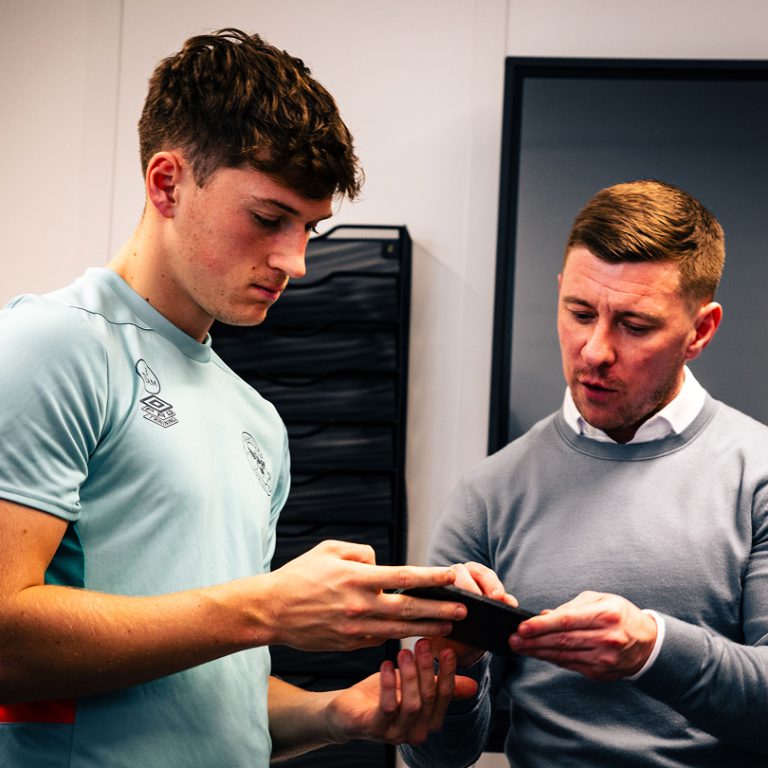 Jack Mackreth, Co-founder of Kyzen Sports showing something on his phone to a man in a football kit