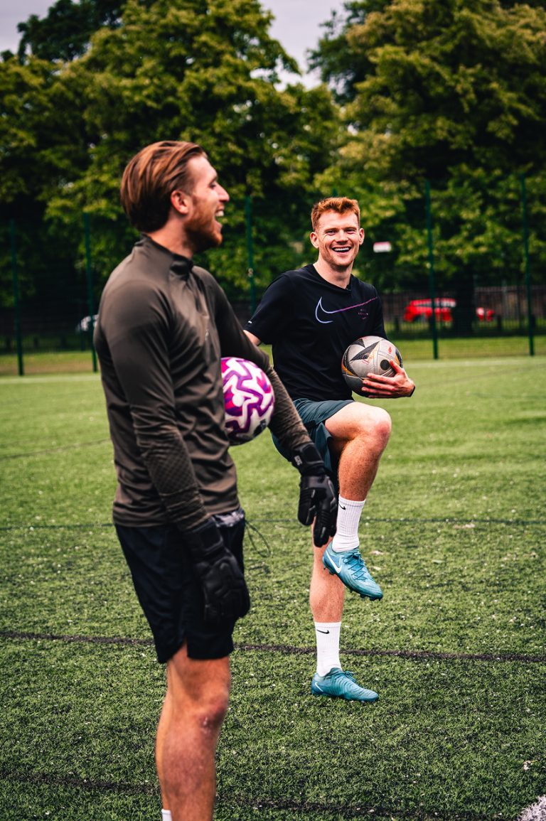 2 men in football gear, each holding a football and laughing