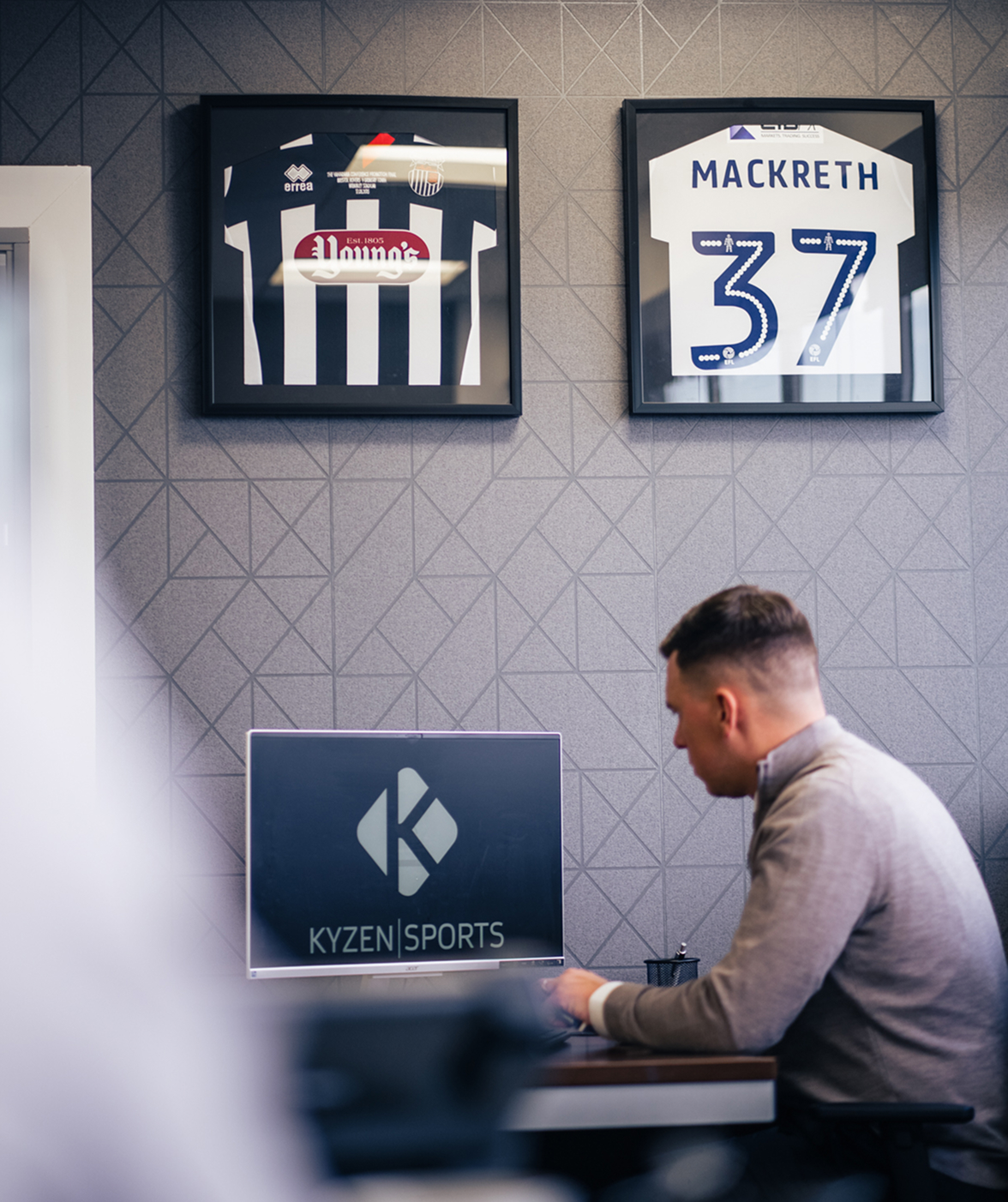 A man sat down in front of a monitor with 2 framed football shirts on the wall behind him